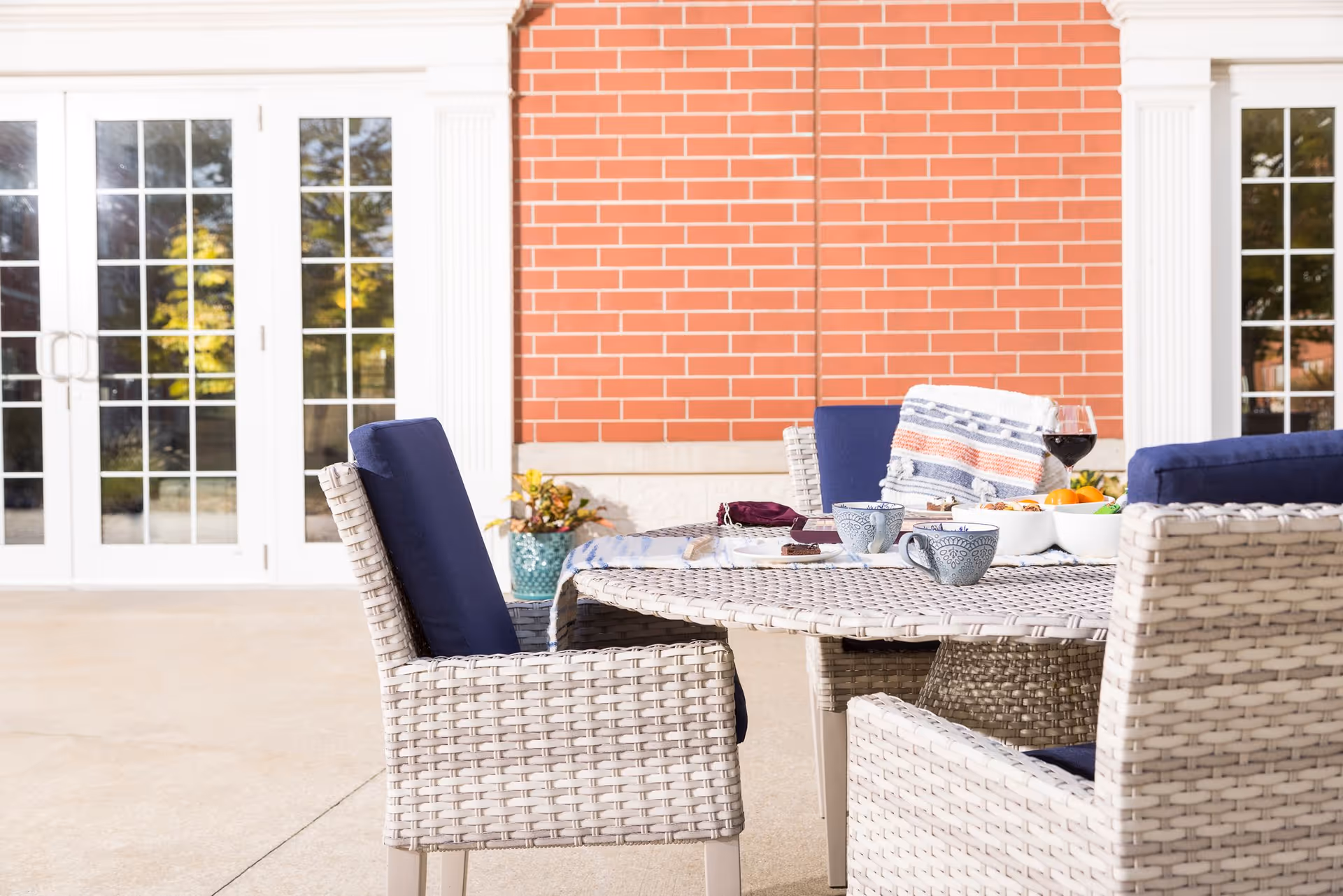 Outdoor patio area with a round wicker table and four wicker chairs with blue cushions. On the table are two blue patterned mugs, a bowl of fruit, a glass of red wine, and a folded blanket. The background shows a brick wall and white-framed glass doors.