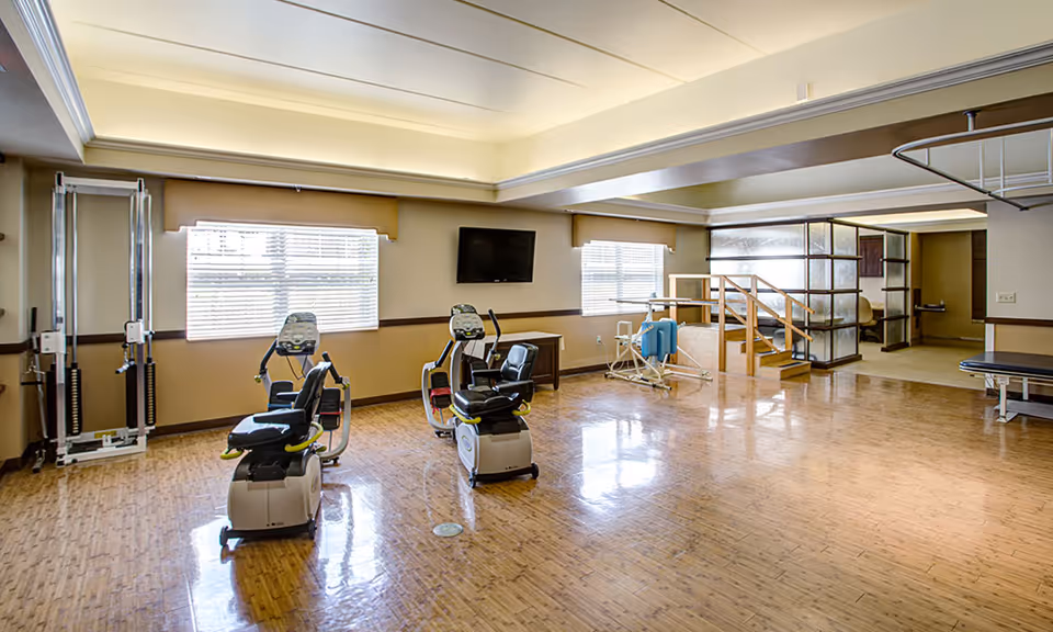 A spacious rehabilitation room with exercise equipment including two recumbent stationary bikes, a weight machine, and parallel bars for physical therapy. The room has large windows with blinds, a wall-mounted TV, wooden flooring, and a small set of stairs with handrails leading to another area.