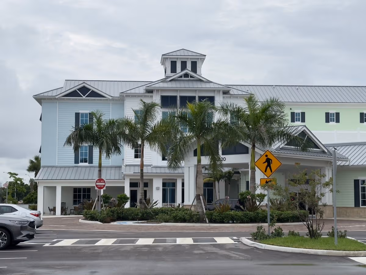 Front exterior of a multi-story coastal-style senior living building with palm trees, a covered entrance, and a parking/driveway area.