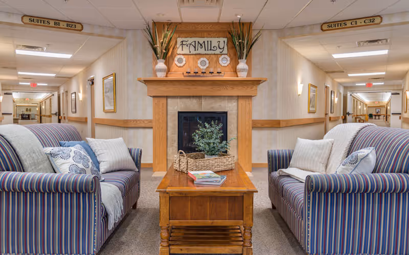 Seating area in a senior living facility with two striped sofas facing a wooden coffee table and a fireplace mantel labeled 'FAMILY', flanked by hallways.