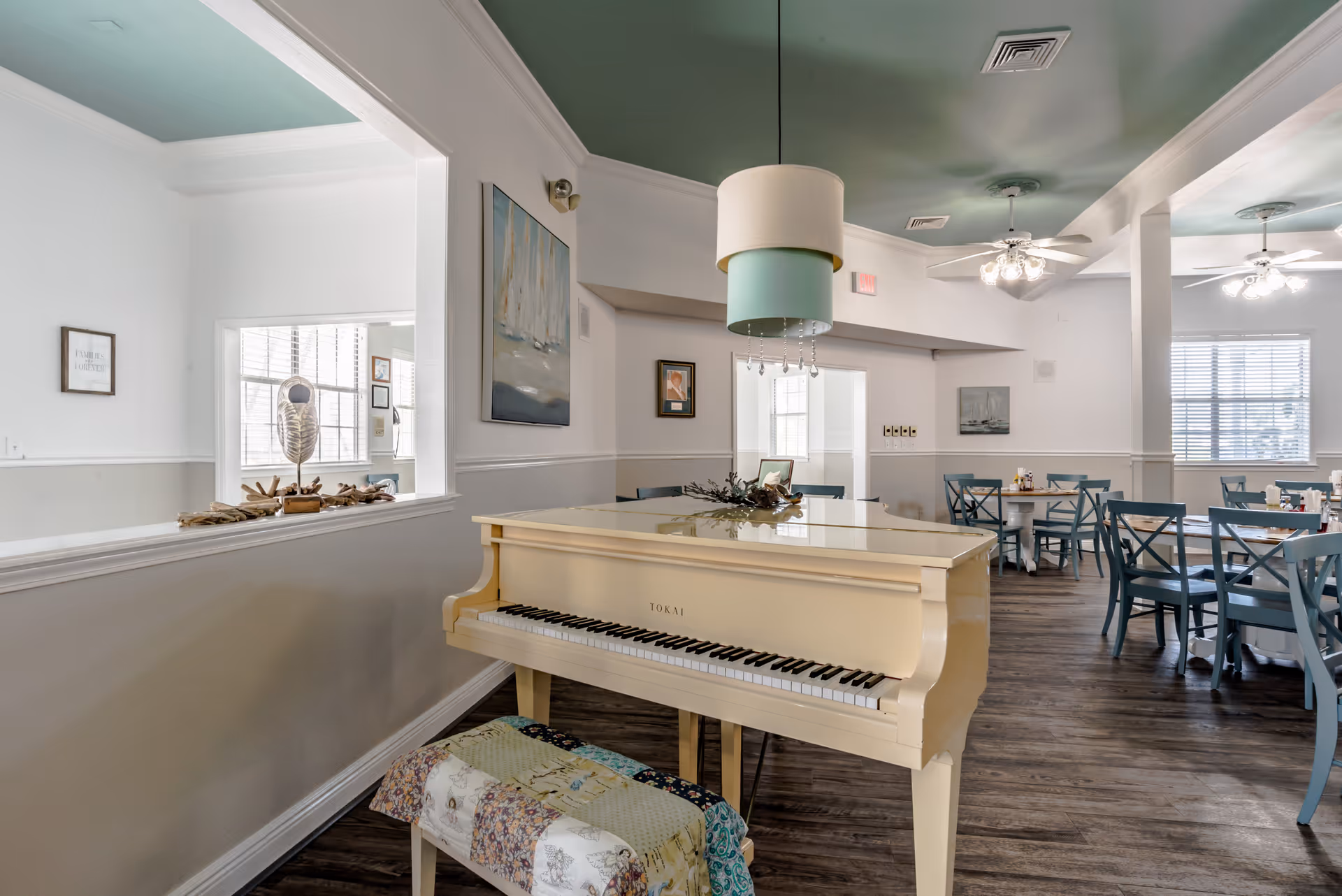 Interior view of a senior living facility dining area with a cream-colored grand piano in the foreground. The piano bench is covered with a patchwork quilt. The room features wooden flooring, light blue chairs around dining tables, ceiling fans with lights, and soft green and white walls. Decorative paintings and a large window letting in natural light are visible.