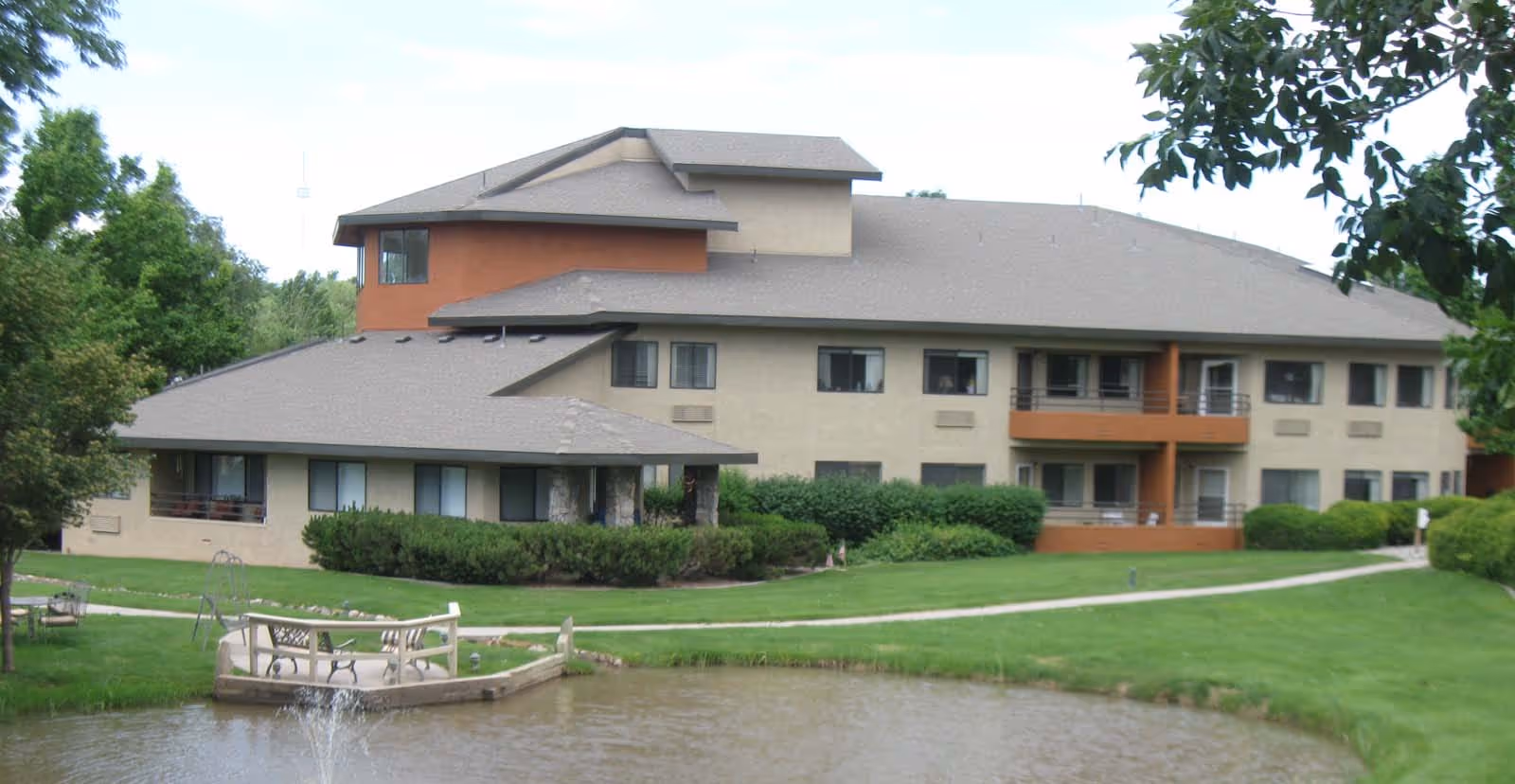 Exterior view of a multi-story senior living facility building with beige and orange walls, surrounded by green lawns, bushes, trees, and a small pond with a fountain in the foreground. There is a paved walkway leading to the building and a small wooden deck with benches near the pond.