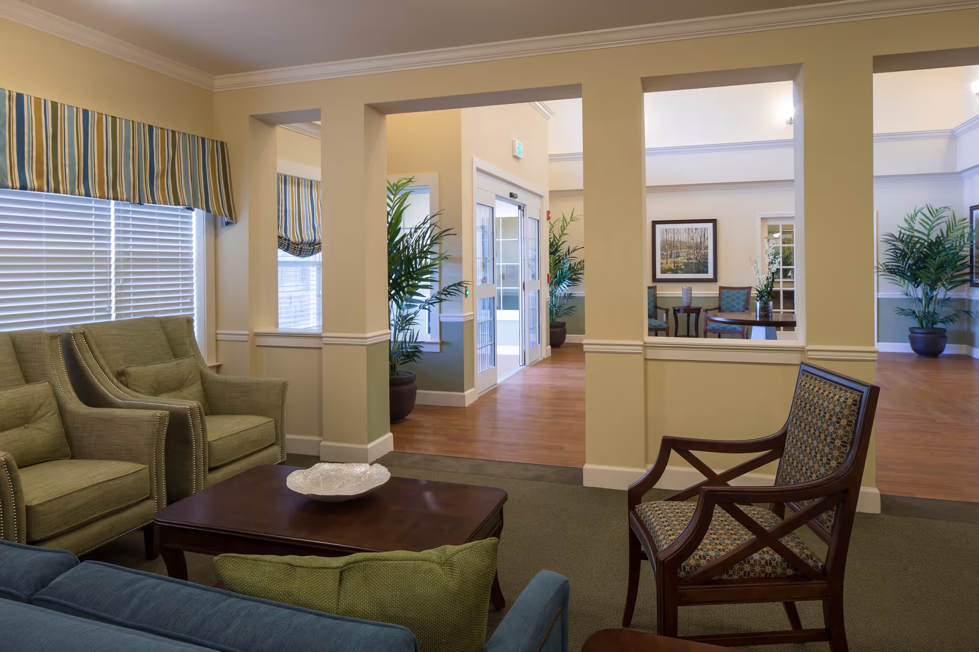 Seating area in a senior living facility lobby with upholstered chairs, a coffee table, and potted plants.