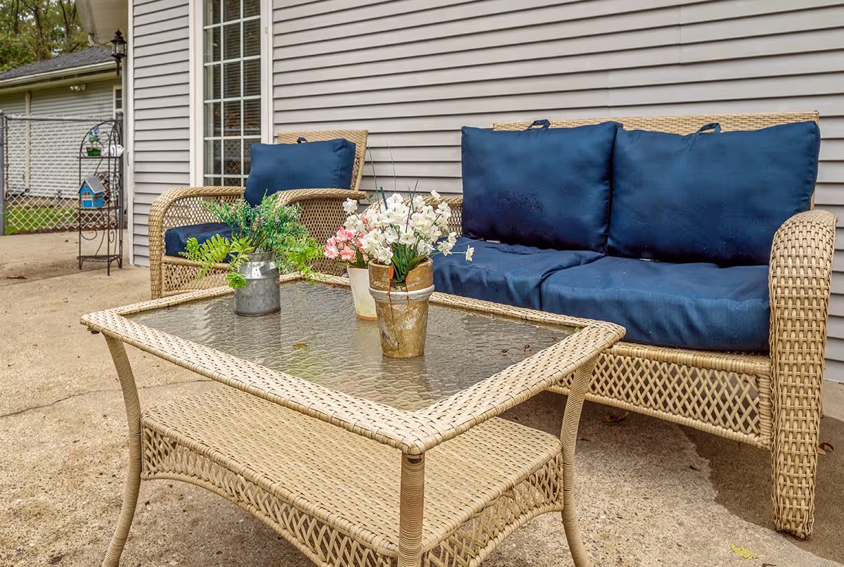 Outdoor patio area with wicker furniture including a loveseat and chair with blue cushions, and a glass-top wicker coffee table with potted plants and flowers. The setting is next to a building with beige siding and a window.