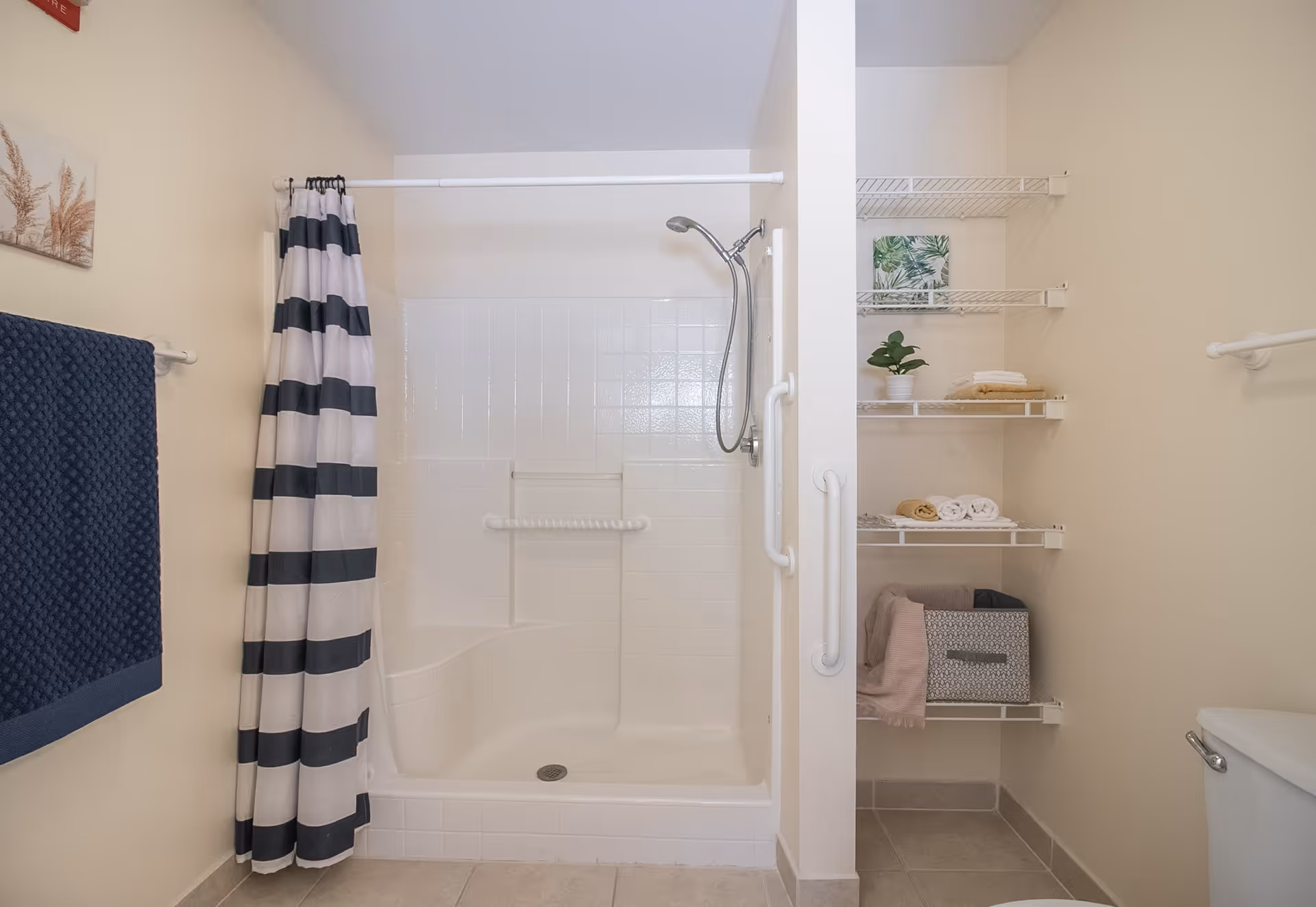 A clean bathroom featuring a walk-in shower with white tiled walls and a striped shower curtain. There are grab bars installed inside and outside the shower for safety. To the right of the shower, there are white wire shelves holding towels, a small plant, and a storage basket. A blue towel hangs on a rack on the left wall, and part of a toilet is visible on the right side of the image.