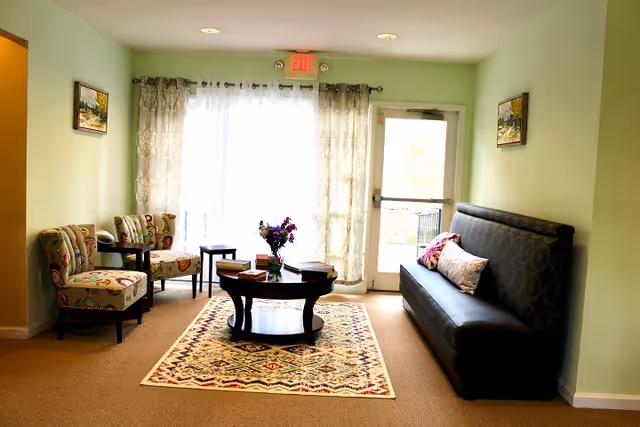 A cozy living room area with light green walls, a patterned rug on the floor, a round wooden coffee table with a vase of flowers and books, two patterned armchairs with a small side table between them on the left, and a dark cushioned bench with pillows on the right. Sheer curtains cover a large window and a glass door leading outside, with an exit sign above the door.