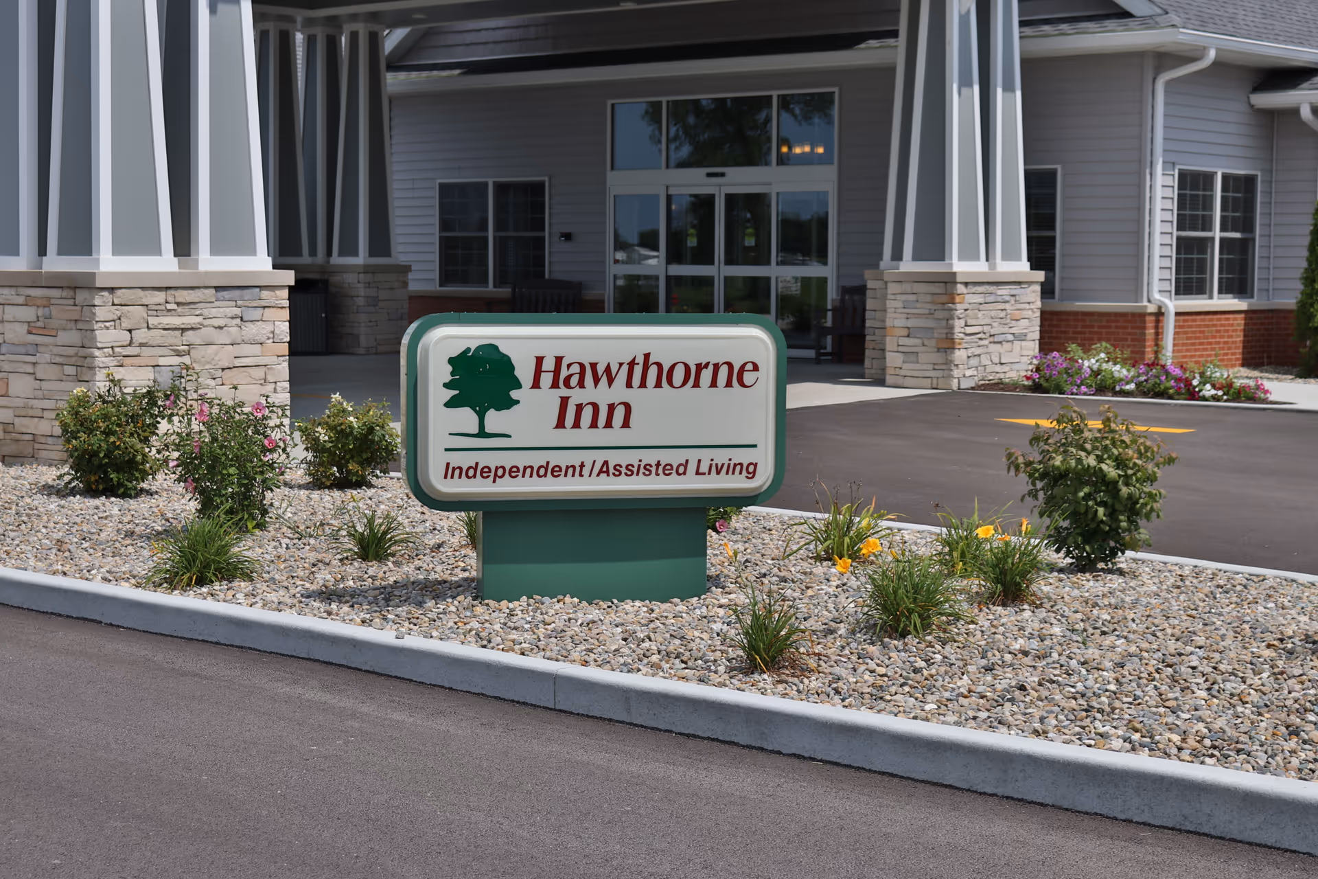 Entrance of a senior living facility with a sign reading 'Hawthorne Inn Independent / Assisted Living' surrounded by small plants and landscaping rocks, with the building entrance visible in the background.