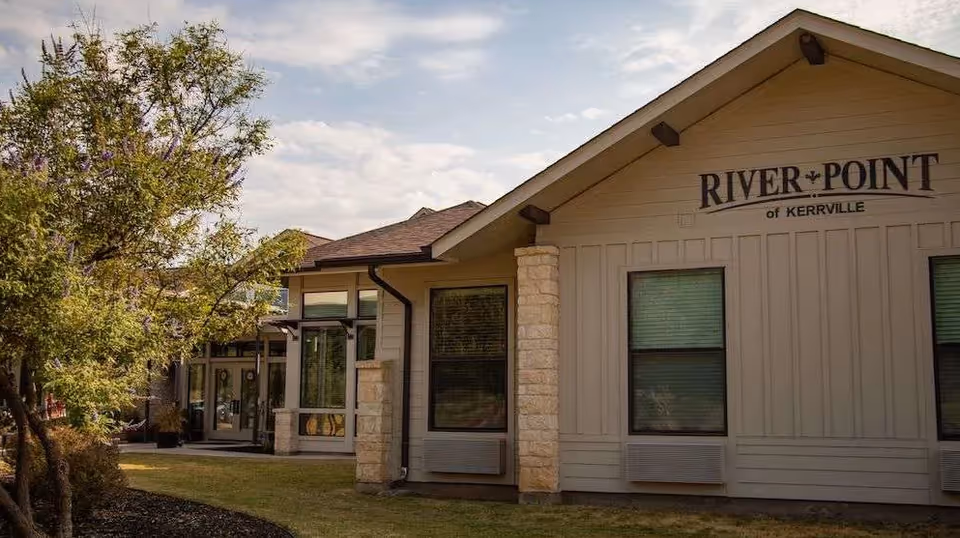 Exterior view of a single-story building with beige siding and stone accents, featuring two windows with air conditioning units below them. The building has a sign that reads 'RIVER POINT of KERRVILLE' near the roof peak. There is a tree and some landscaping in the foreground under a partly cloudy sky.