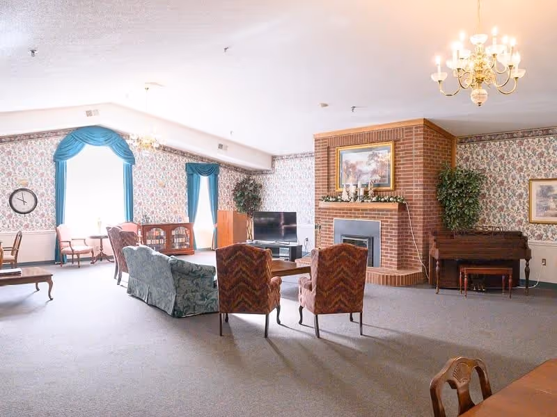 A spacious living room with floral wallpaper and blue curtains. The room features a brick fireplace with a framed painting above it, two patterned armchairs, a green sofa, a wooden coffee table, a flat-screen TV on a stand, a piano, and a chandelier hanging from the ceiling. There are also additional seating areas near the windows with natural light coming in.