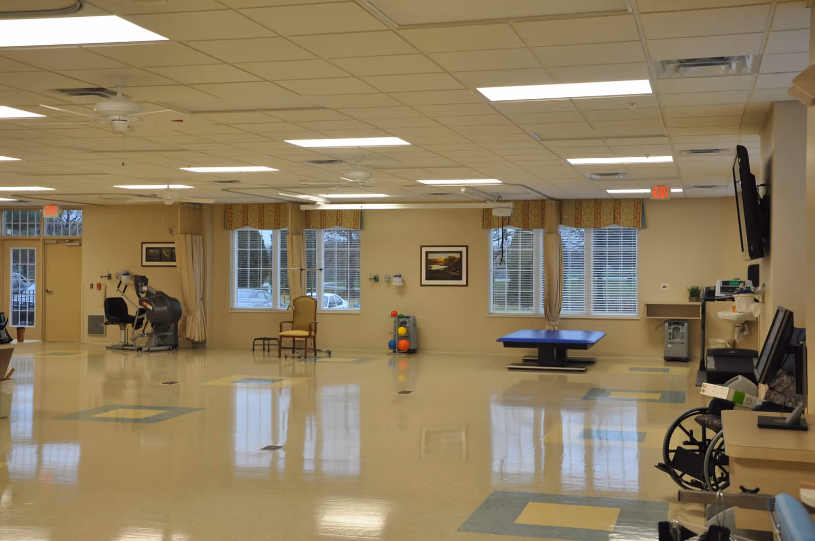 A spacious rehabilitation room with large windows, exercise equipment, a wheelchair, a chair, a therapy table, and medical supplies. The room has a tiled floor with geometric patterns and ceiling fans with fluorescent lighting.