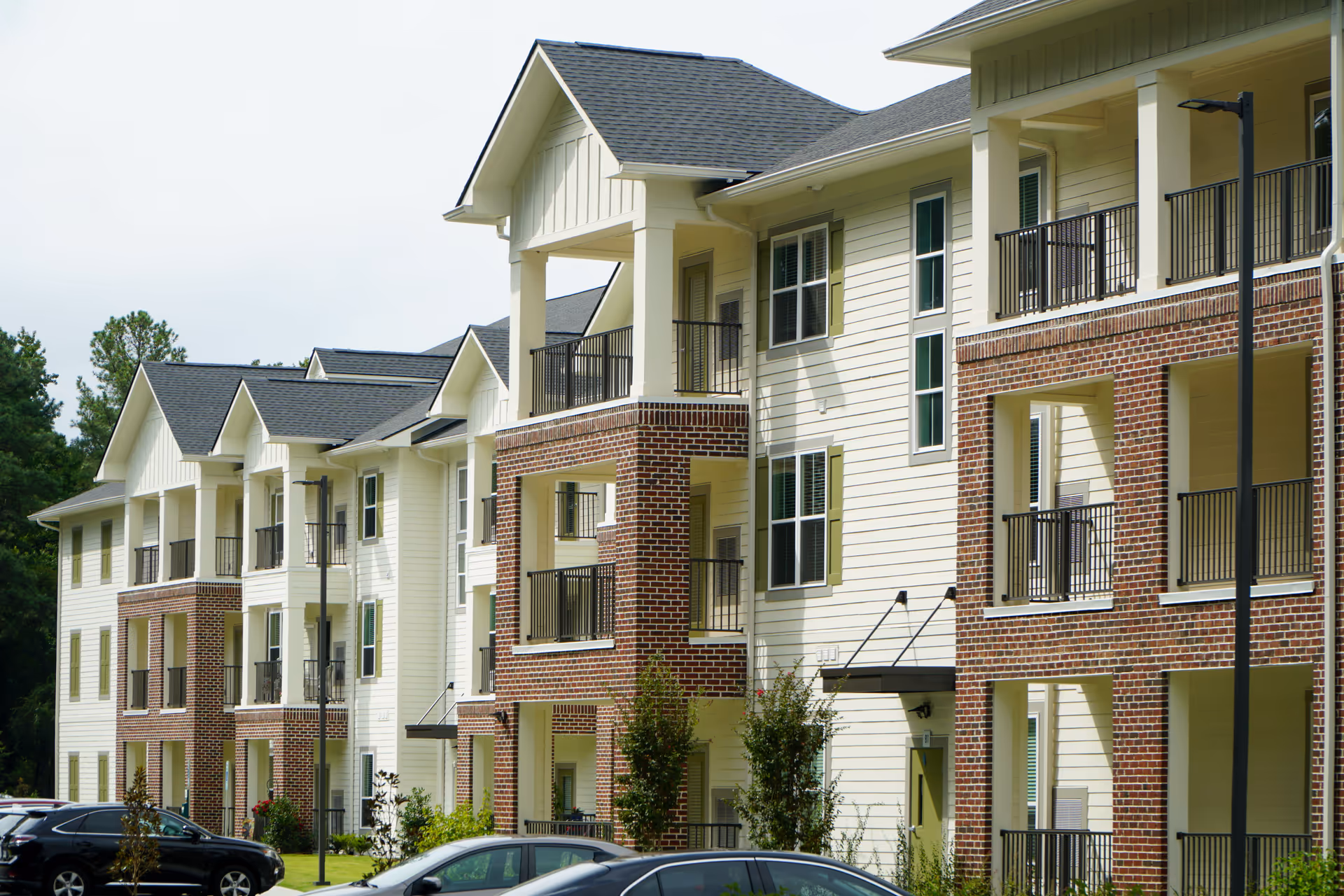 Exterior view of a multi-story residential building with white siding and red brick accents, featuring balconies and a parking area with several cars in front.