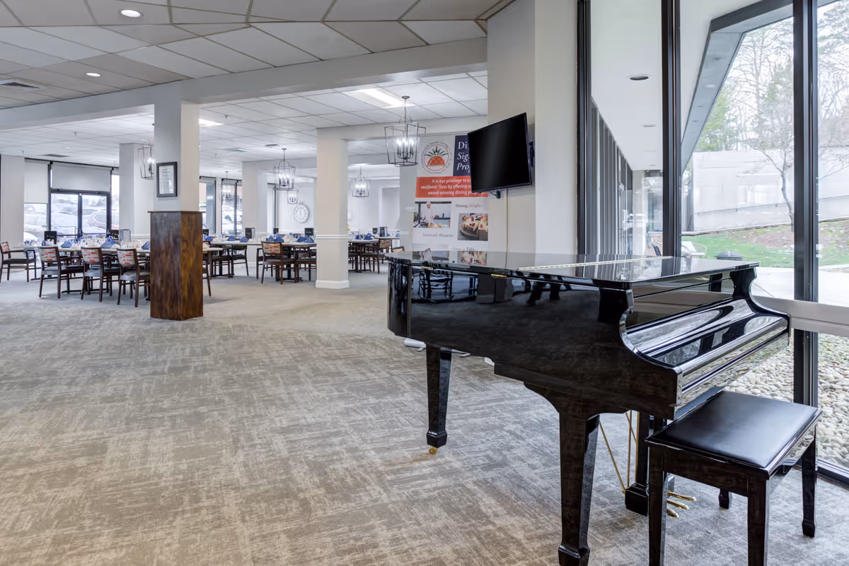 Interior view of a senior living facility dining room with multiple tables and chairs set for meals. A black grand piano with a matching bench is positioned near large windows overlooking an outdoor area. The room has carpeted floors, white walls, and ceiling lights with decorative fixtures. A wall-mounted TV and a sign about the dining program are visible.