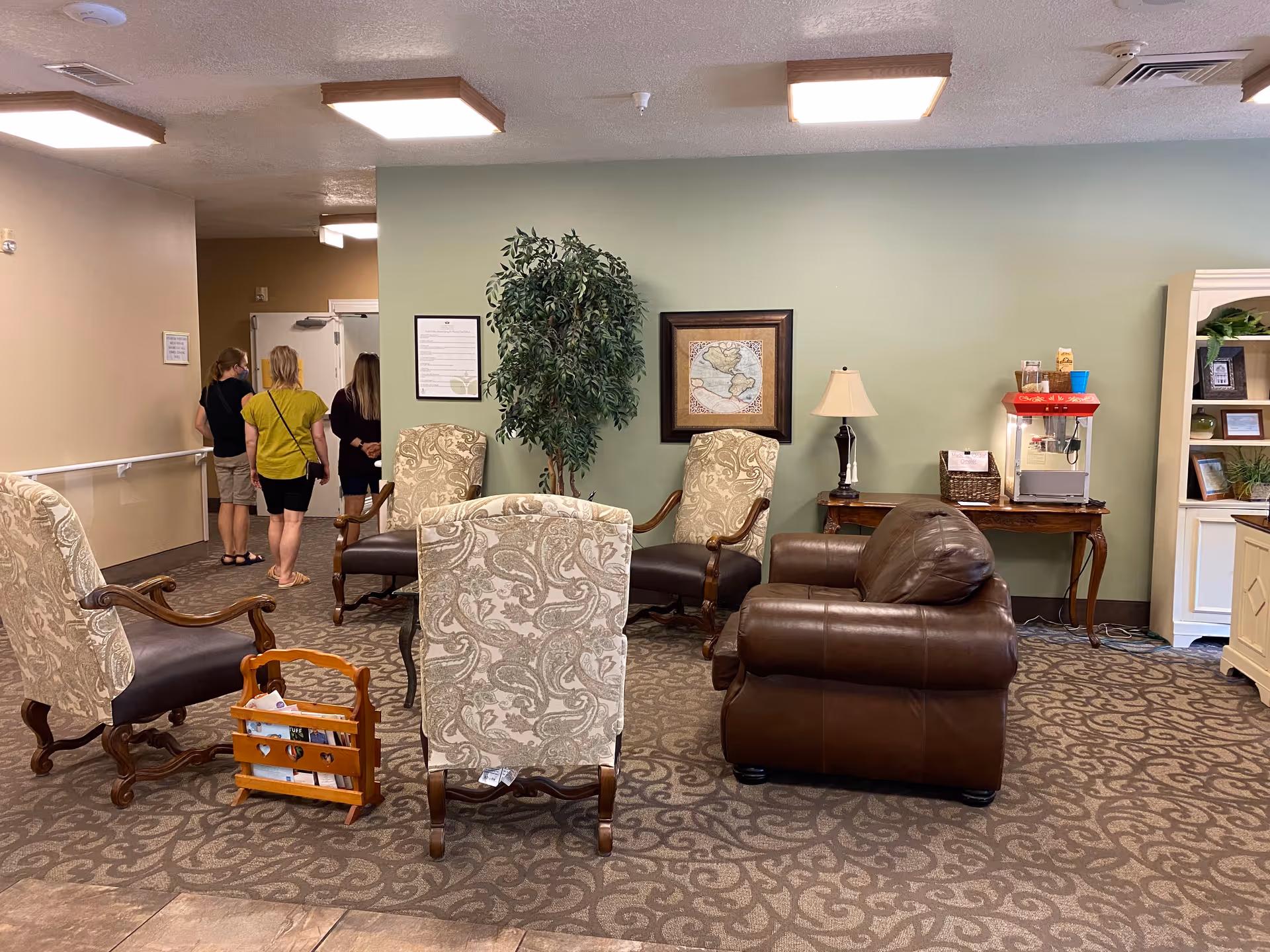 A cozy seating area in an assisted living facility with patterned armchairs and a brown leather armchair arranged on a patterned carpet. A small wooden table with a lamp and a popcorn machine is against a light green wall, which also features a framed map and a potted plant. In the background, three people are standing near a hallway.