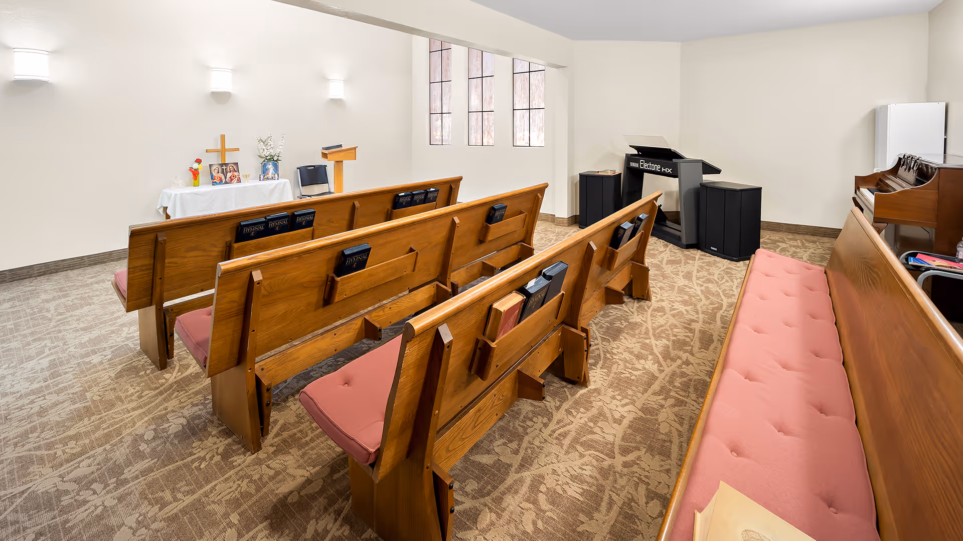Interior view of a small chapel room with wooden pews featuring pink cushions, a lectern, a cross on a table with flowers and religious pictures, a keyboard, and a piano. The room has beige walls, carpeted floor, and windows letting in natural light.