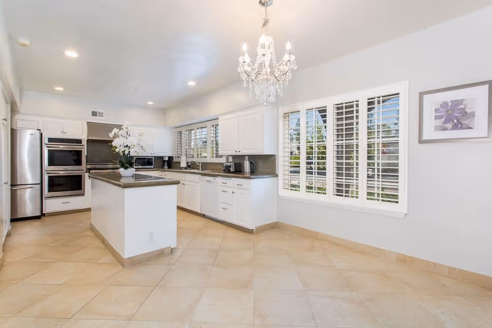 Bright and spacious kitchen with white cabinets, a central island with a potted orchid, stainless steel refrigerator and double oven, large windows with white shutters, beige tiled floor, and a chandelier hanging from the ceiling.