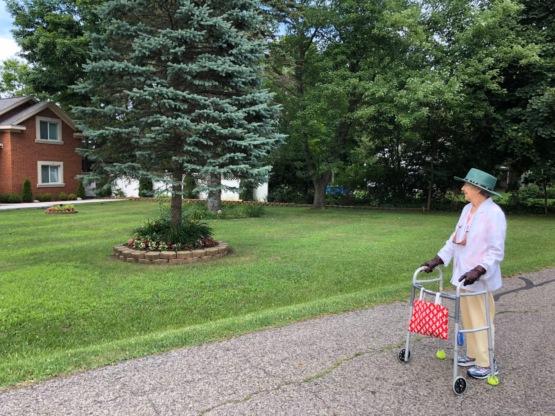 An elderly woman wearing a green hat, white jacket, beige pants, and gloves is walking with a walker on a paved path in a grassy outdoor area. There are trees and a brick building in the background.