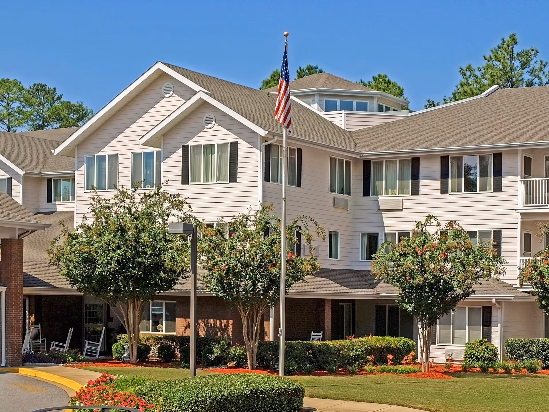 Exterior view of a senior living facility building with beige siding and multiple windows, surrounded by well-maintained landscaping including trees, bushes, and flowers. An American flag is displayed on a flagpole in front of the building under a clear blue sky.