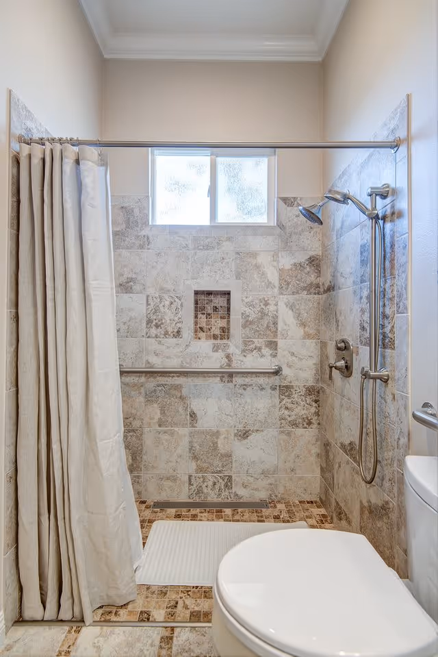 A bathroom shower area with beige and brown tiled walls and floor, a silver showerhead with an adjustable handheld attachment, a grab bar on the wall, a white shower curtain partially drawn to the left, a small frosted window above the shower, and a white toilet partially visible on the right.