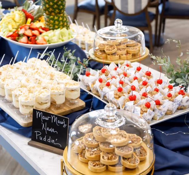 A dessert buffet table featuring individual servings of banana pudding with spoons, mini pecan pies under glass covers, and cupcakes topped with cherries. A plate of fresh fruit including pineapple, strawberries, and melon is also visible in the background. The table is covered with a dark blue cloth and decorated with greenery.