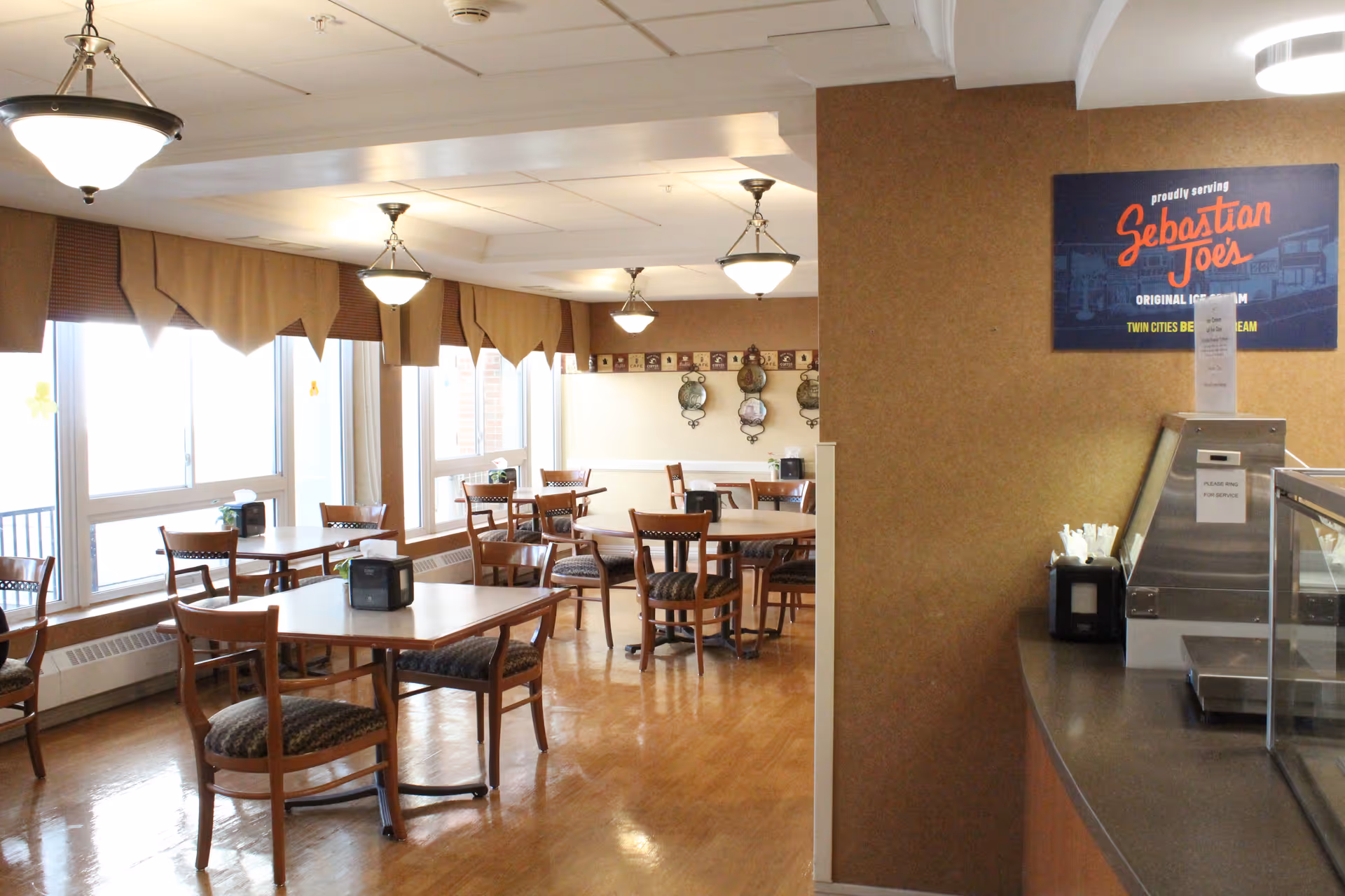 A dining area with several wooden tables and chairs arranged neatly. Large windows with valance curtains allow natural light to fill the room. On the right side, there is a counter with a sign above it that reads 'Sebastian Joe's Original Ice Cream Twin Cities Best Ice Cream'. The floor is polished wood, and the ceiling has multiple hanging light fixtures.