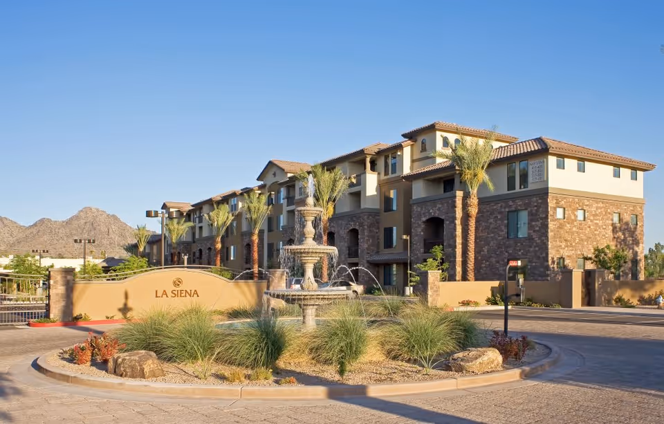 Exterior view of La Siena senior living facility with a multi-story building, palm trees, a decorative fountain, and desert landscaping under a clear blue sky.