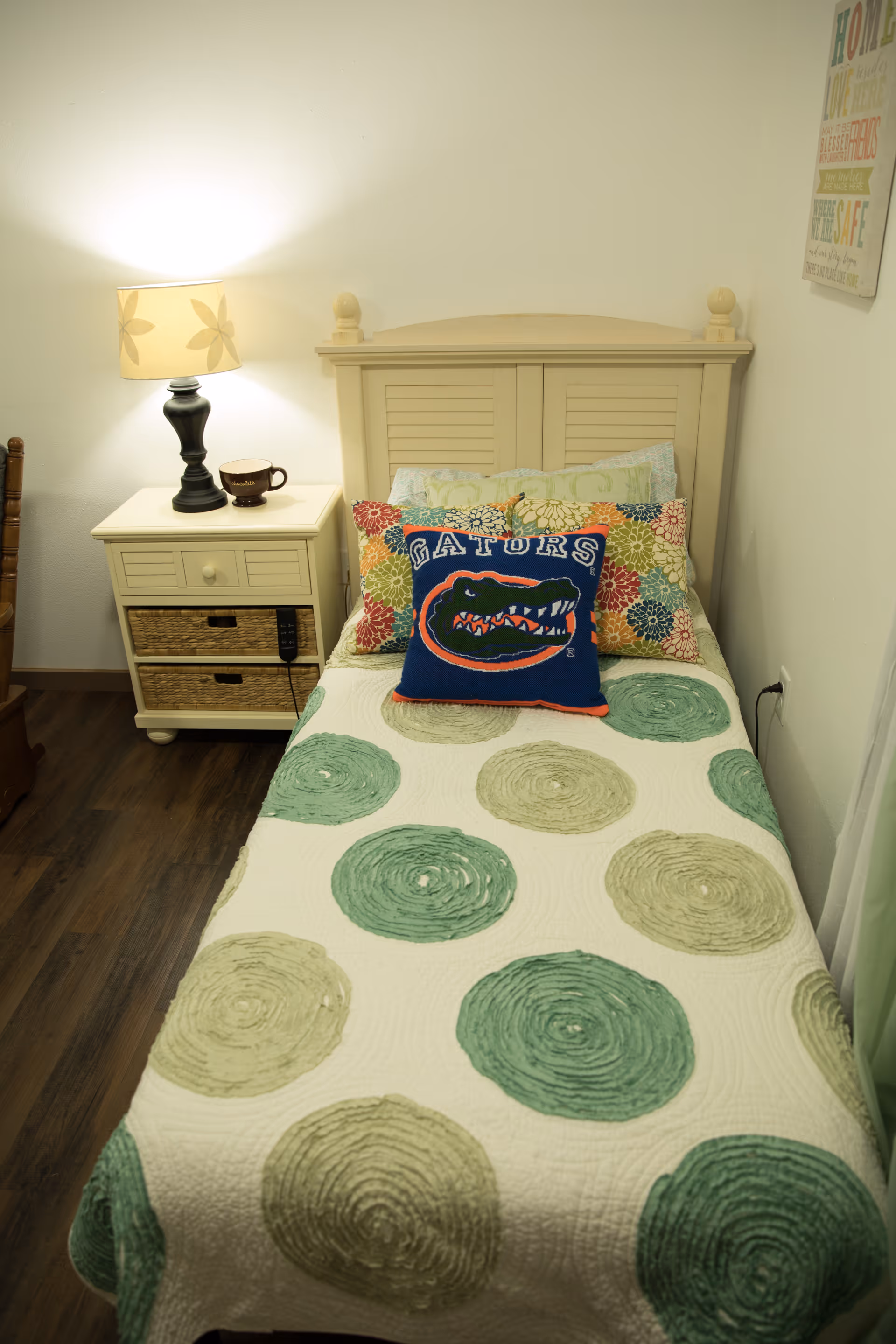 A small bedroom with a single bed featuring a quilt with green and beige circular patterns. The bed has multiple pillows, including a decorative pillow with the Florida Gators logo. Next to the bed is a cream-colored nightstand with two wicker drawers, a lamp with a floral lampshade, and a coffee mug. The room has wooden flooring and light-colored walls with a framed inspirational quote hanging on the right wall.