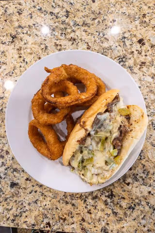 A white plate on a granite countertop holding a cheesesteak sandwich with melted cheese and green peppers, accompanied by several golden-brown onion rings.
