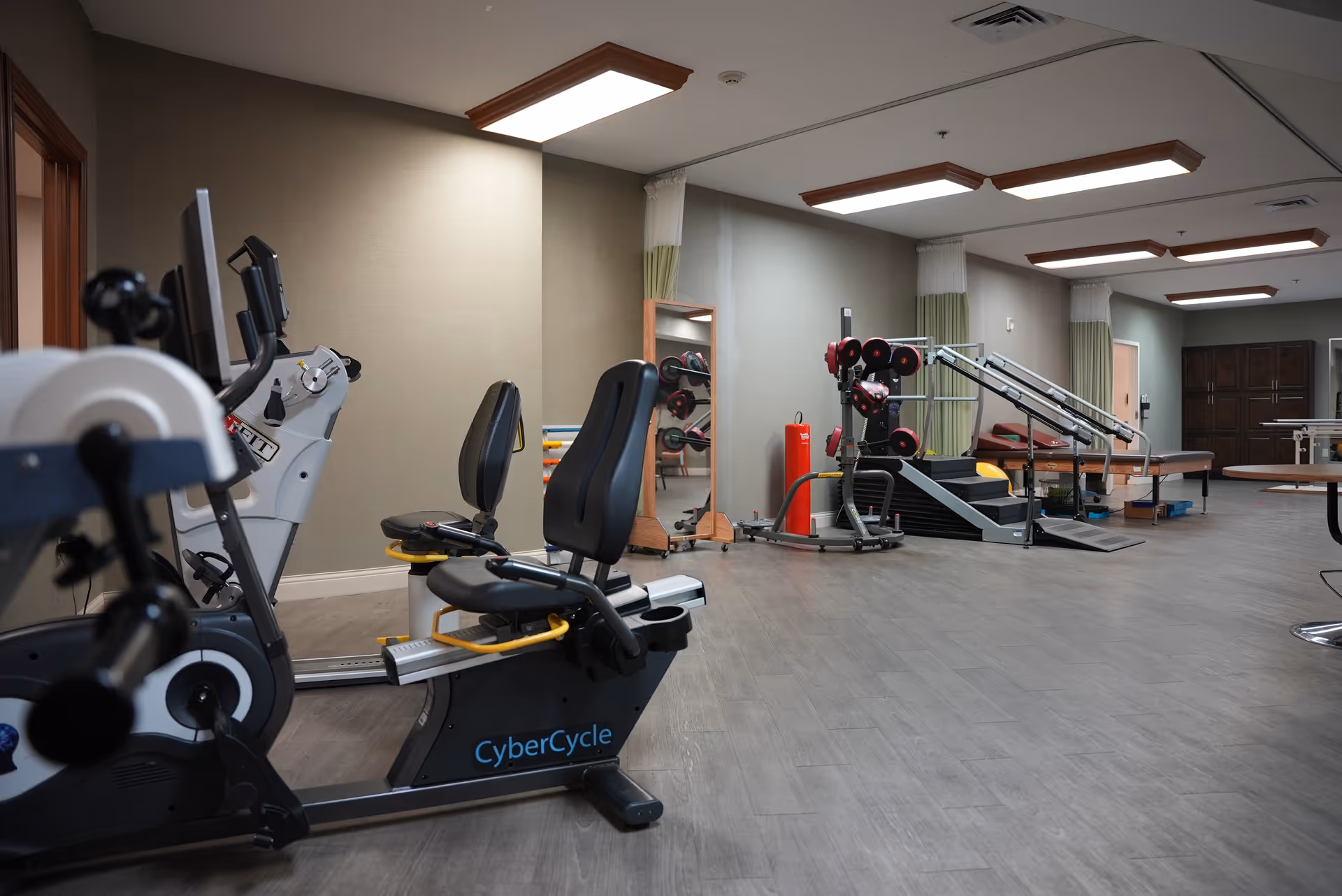 Interior view of a fitness room in a senior living facility with exercise equipment including a CyberCycle recumbent bike, weight machines, and a therapy table. The room has wood-like flooring, beige walls, ceiling lights, and a large mirror on one wall.