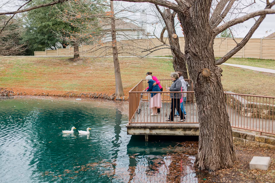 Three elderly women standing on a wooden deck overlooking a pond with two white ducks swimming. One woman is using a walker, and the other two are standing nearby. The scene is outdoors with trees and a grassy area in the background.