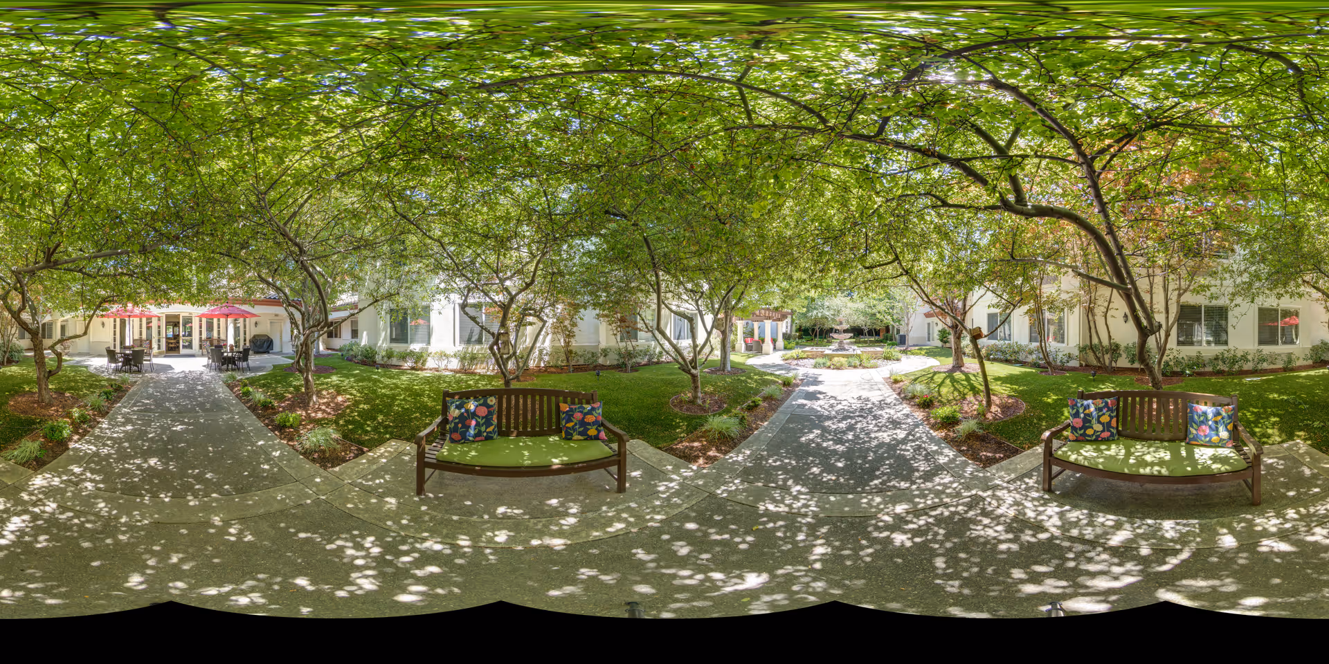 A shaded outdoor courtyard area with two wooden benches featuring green cushions and colorful pillows. The courtyard is surrounded by trees with dappled sunlight filtering through the leaves, and there are pathways leading to a building entrance with red umbrellas and outdoor seating.