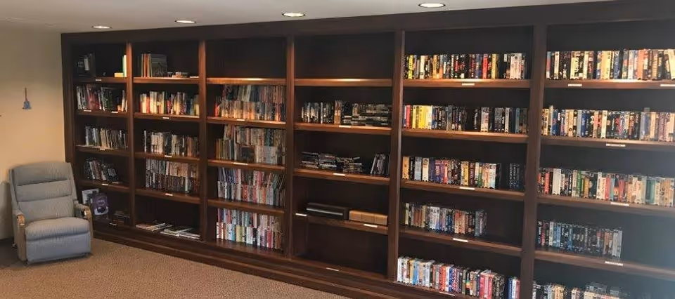 A cozy room with a large wooden bookshelf filled with books and DVDs, and a single cushioned armchair positioned to the left side of the bookshelf.