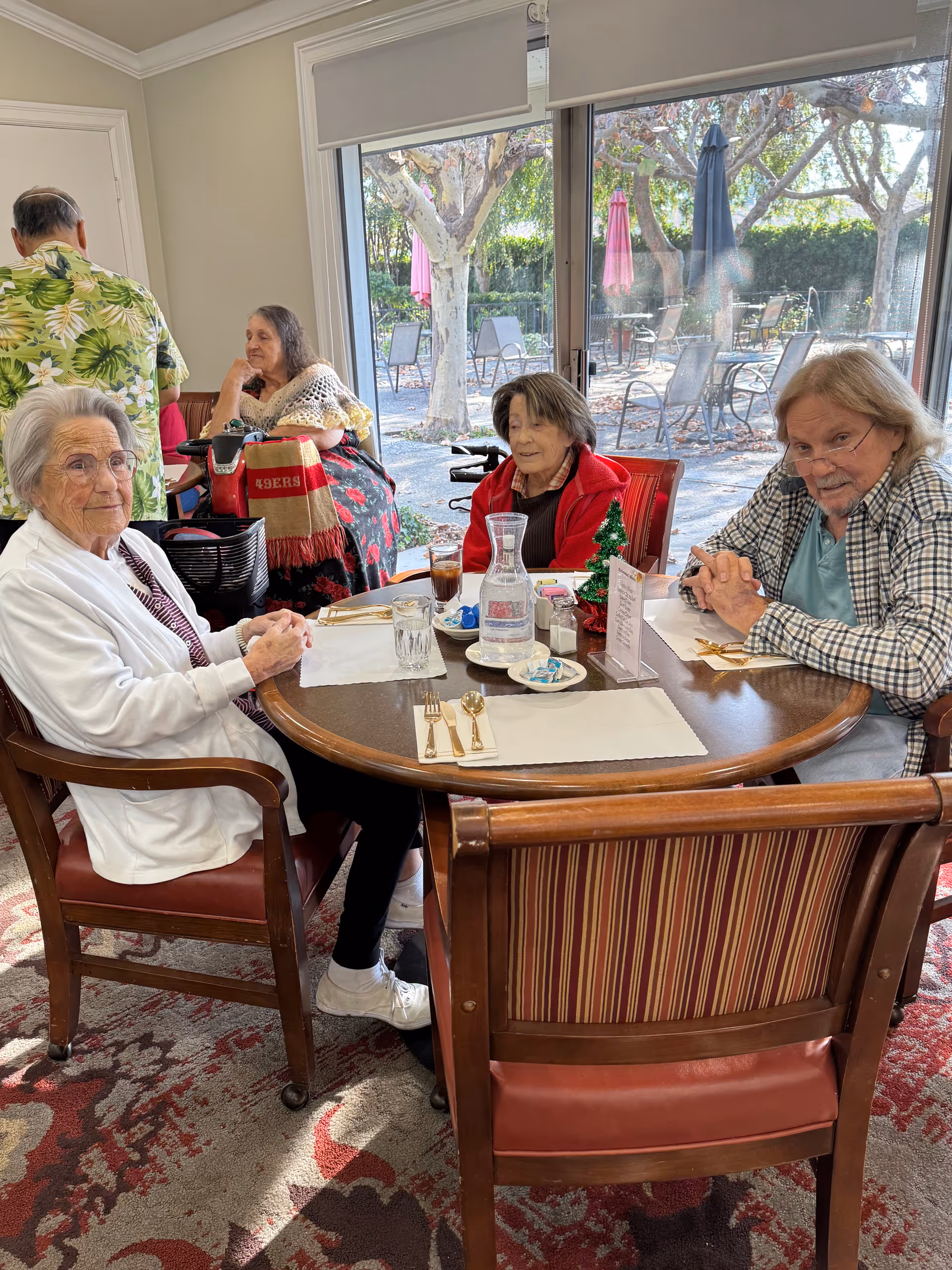 Four elderly individuals sitting around a dining table inside a room with large windows showing an outdoor patio with tables, chairs, and umbrellas. The table is set with placemats, utensils, glasses, a water pitcher, and a small Christmas tree decoration. One person is in a wheelchair with a 49ers blanket, and the others are seated in wooden chairs with red cushions.