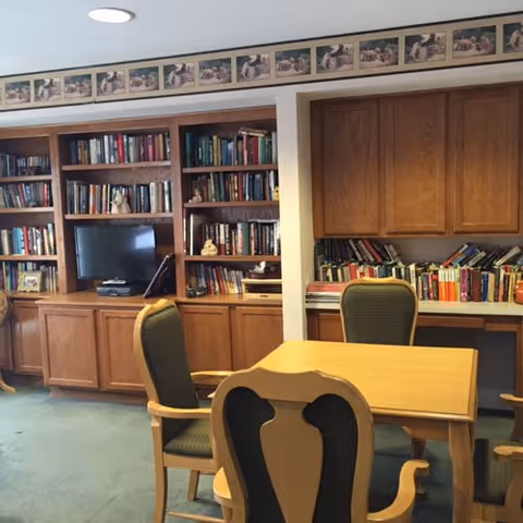 A cozy room with wooden bookshelves filled with books, a small television, and a wooden table surrounded by four cushioned chairs. The room has a green carpet and a decorative border near the ceiling featuring images of people reading.