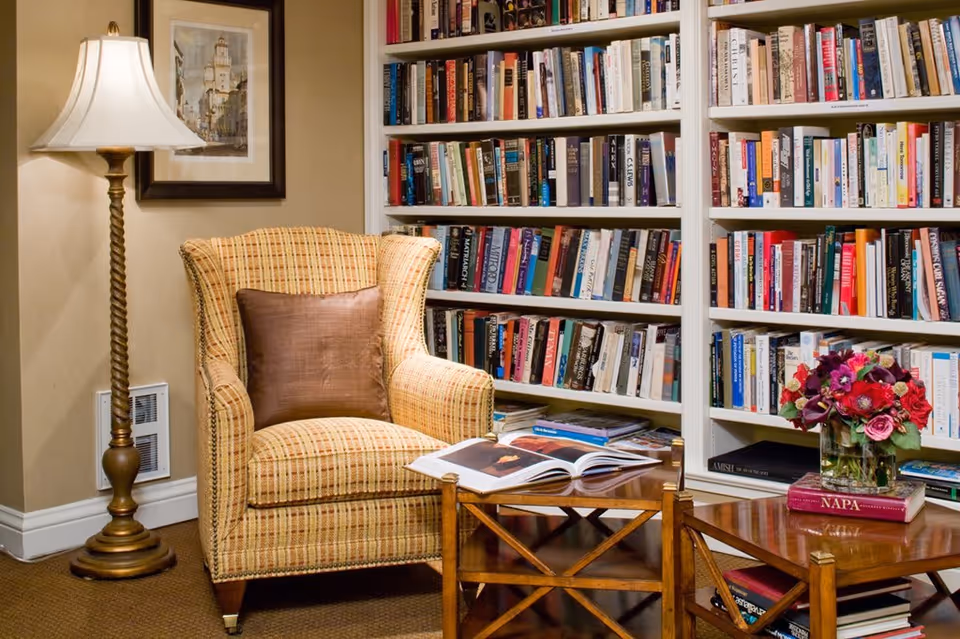 Cozy reading nook featuring a cushioned armchair with a brown pillow, a wooden floor lamp with a white lampshade, and a wooden coffee table with an open book and a vase of flowers. Behind the chair is a large white bookshelf filled with numerous books.