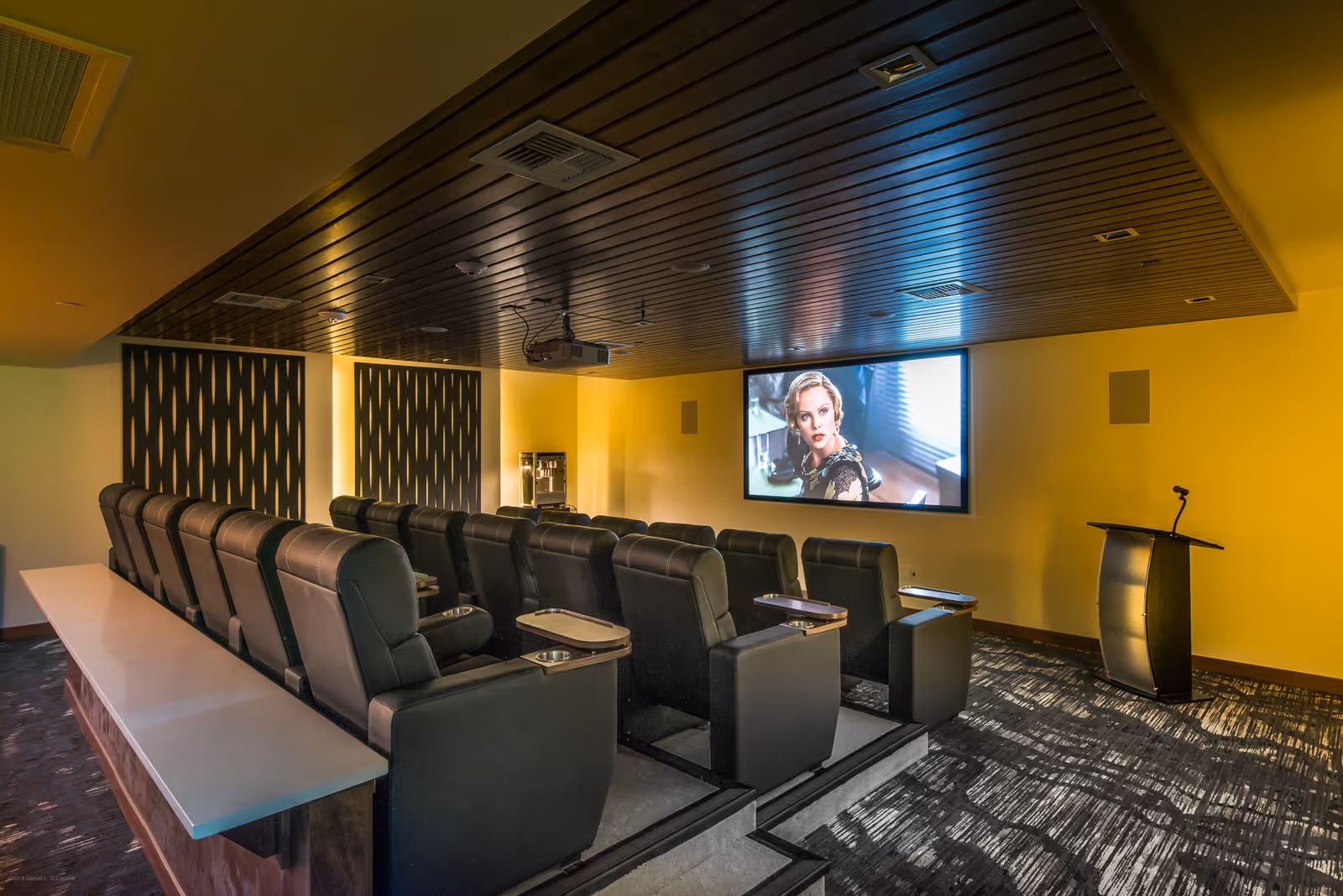 A small theater room with two rows of black leather recliner seats facing a large screen displaying a woman. The room has a dark wooden ceiling with recessed lighting, patterned carpet, and a podium with a microphone on the right side.