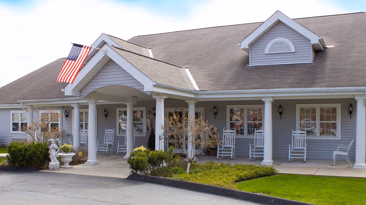 Front entrance of a single-story senior living facility with a covered porch, white columns, rocking chairs, landscaping, and an American flag.