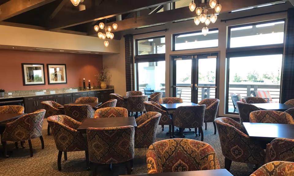 Bright communal dining area with patterned upholstered chairs and square tables, chandeliers, and large windows/doors to an outdoor patio.
