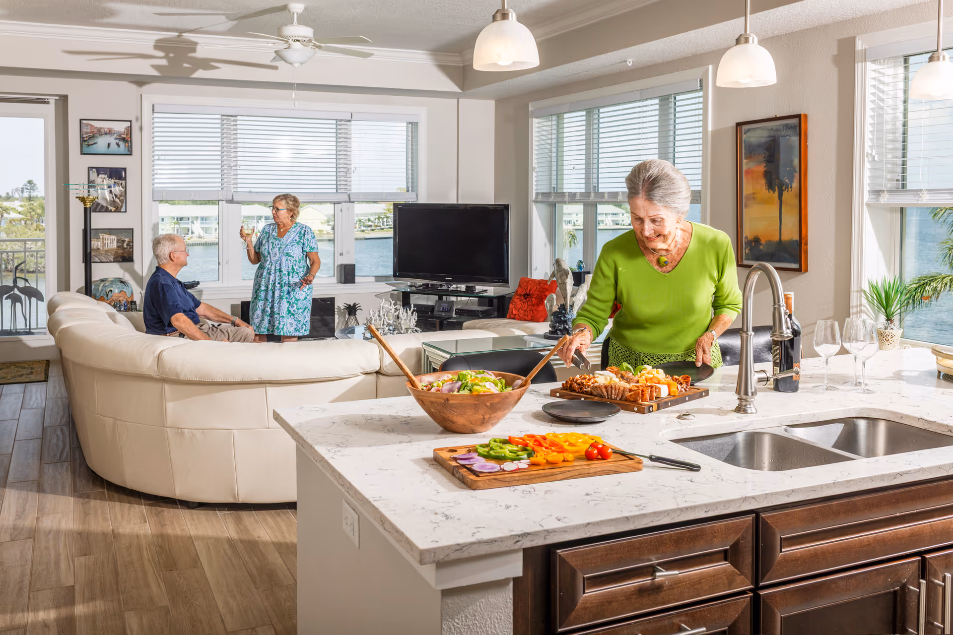 A bright and spacious living area with large windows overlooking a waterfront. In the foreground, an elderly woman in a green top is preparing food on a kitchen island with a cutting board of colorful vegetables and a wooden bowl of salad. In the background, two elderly people are conversing near a curved white sofa. The room features wooden flooring, a ceiling fan, and a TV on a stand.