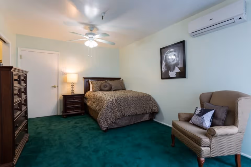A bedroom with green carpet, a bed with patterned bedding, a wooden nightstand with a lamp, a wooden dresser, a beige armchair with a decorative pillow, a ceiling fan with lights, and a framed black and white portrait on the wall.