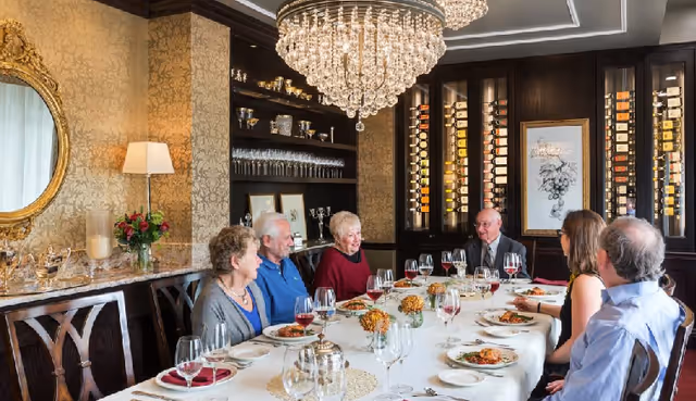 A group of six elderly and middle-aged people sitting around a long dining table in an elegant dining room. The table is set with plates of food, wine glasses, and floral centerpieces. The room features a large crystal chandelier, a wine display cabinet, a round gold-framed mirror, and warm decorative wallpaper.