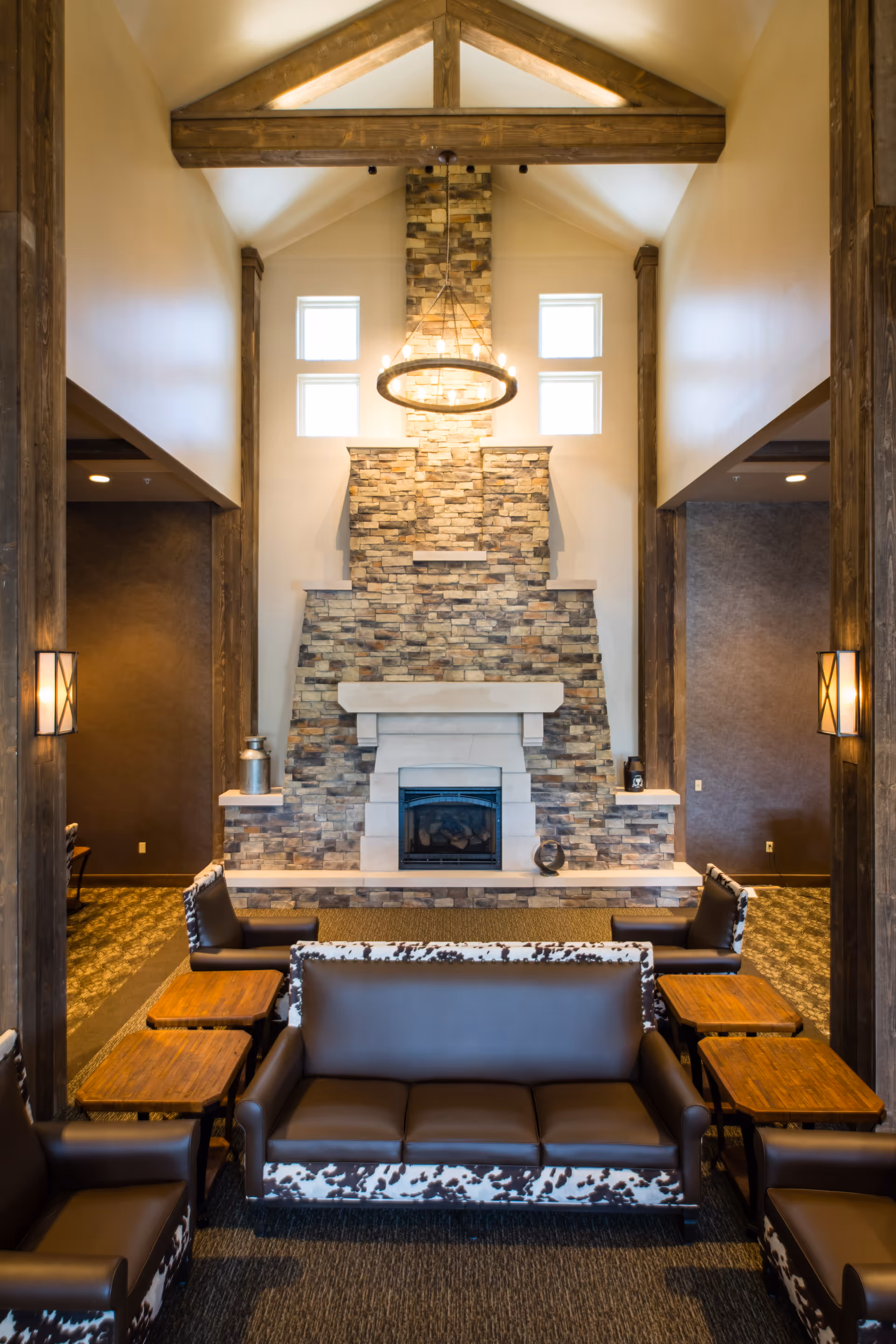 A rustic communal lounge with a tall stone fireplace, chandelier, vaulted ceiling, and leather sofas arranged around side tables.