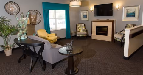 A cozy living room area with a gray sofa, yellow armchair, two patterned chairs, a fireplace with a mounted TV above it, and decorative wall art. There is a large window with teal curtains and a couple of round tables with decorative items on them.