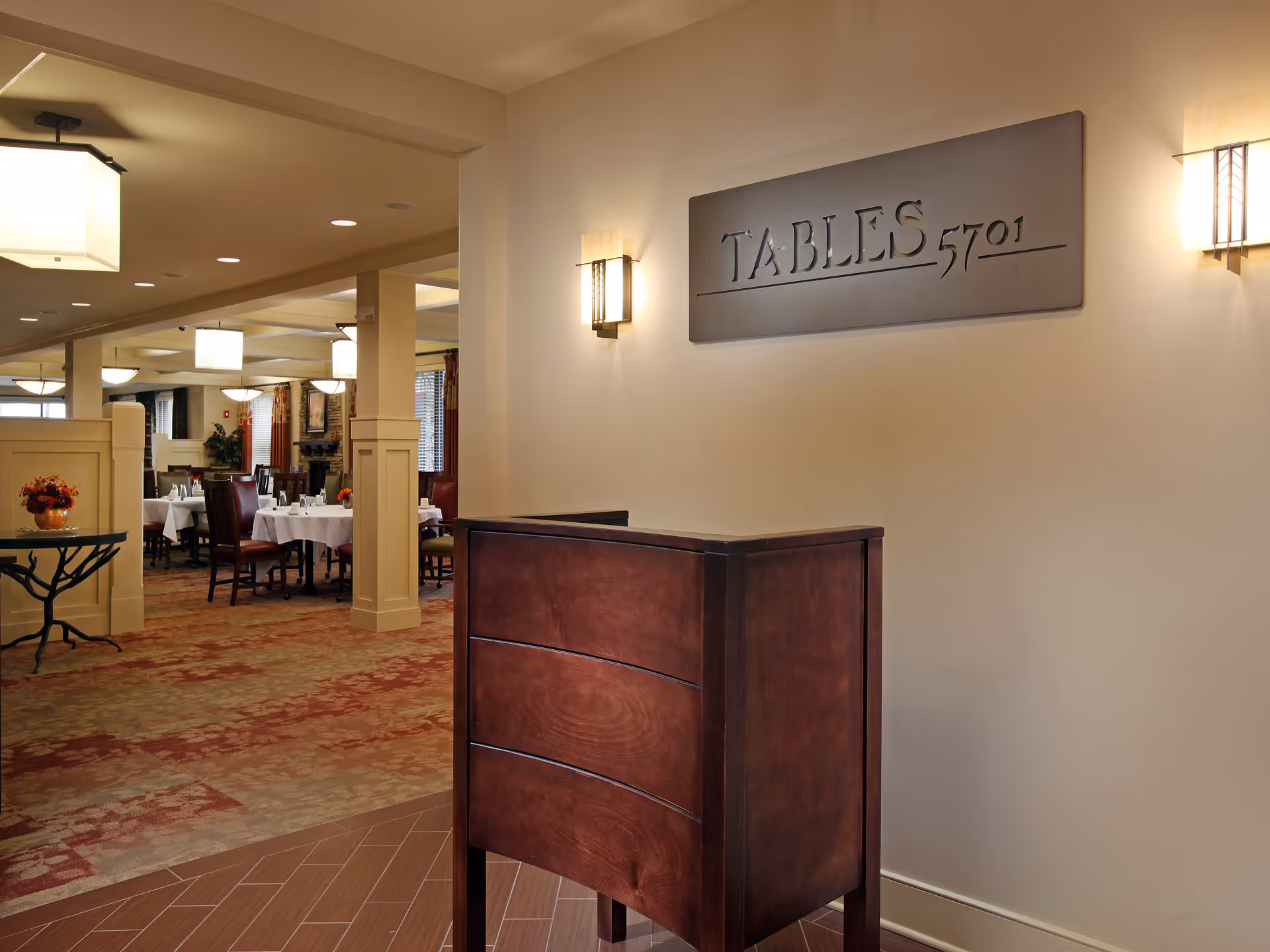 Interior view of a dining area in a senior living facility with a wooden hostess stand in the foreground and tables with white tablecloths and chairs in the background. A sign on the wall reads 'TABLES 5701'. The room is warmly lit with wall sconces and ceiling lights.