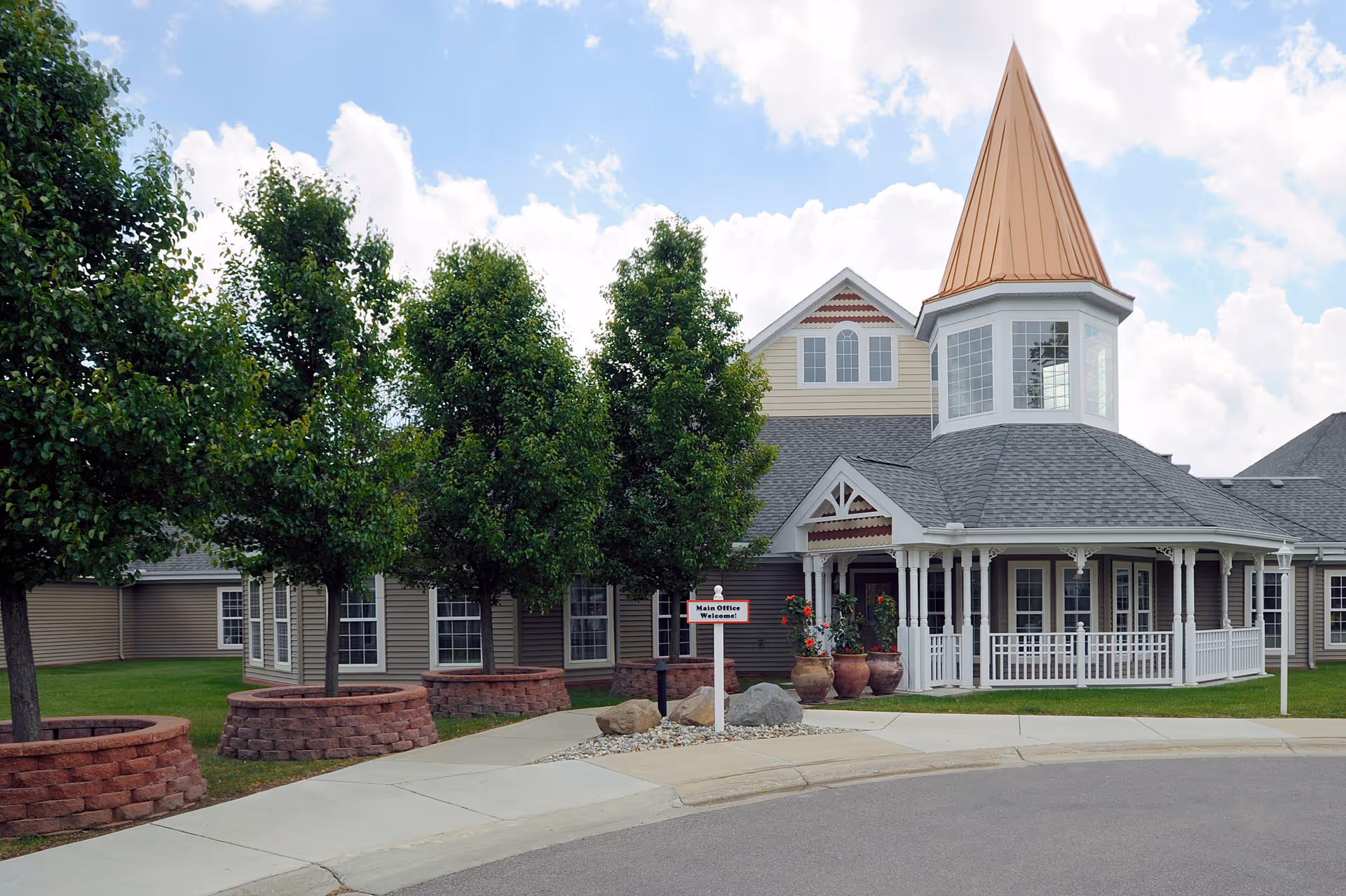 Exterior view of a senior living facility building with a pointed turret roof, multiple windows, a white porch railing, and several trees planted in circular brick planters along a curved sidewalk.