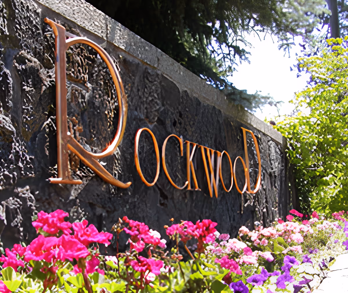 Stone wall with the word 'Rockwood' in large metal letters, surrounded by colorful flowers and greenery under a bright sky.