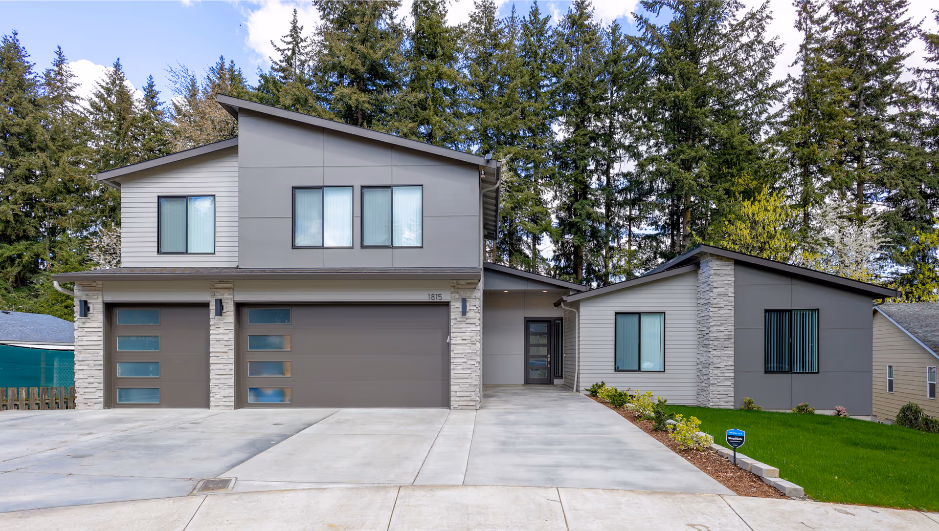 Modern two-story gray house with multiple garage doors, a concrete driveway, and a landscaped lawn in front of tall trees.