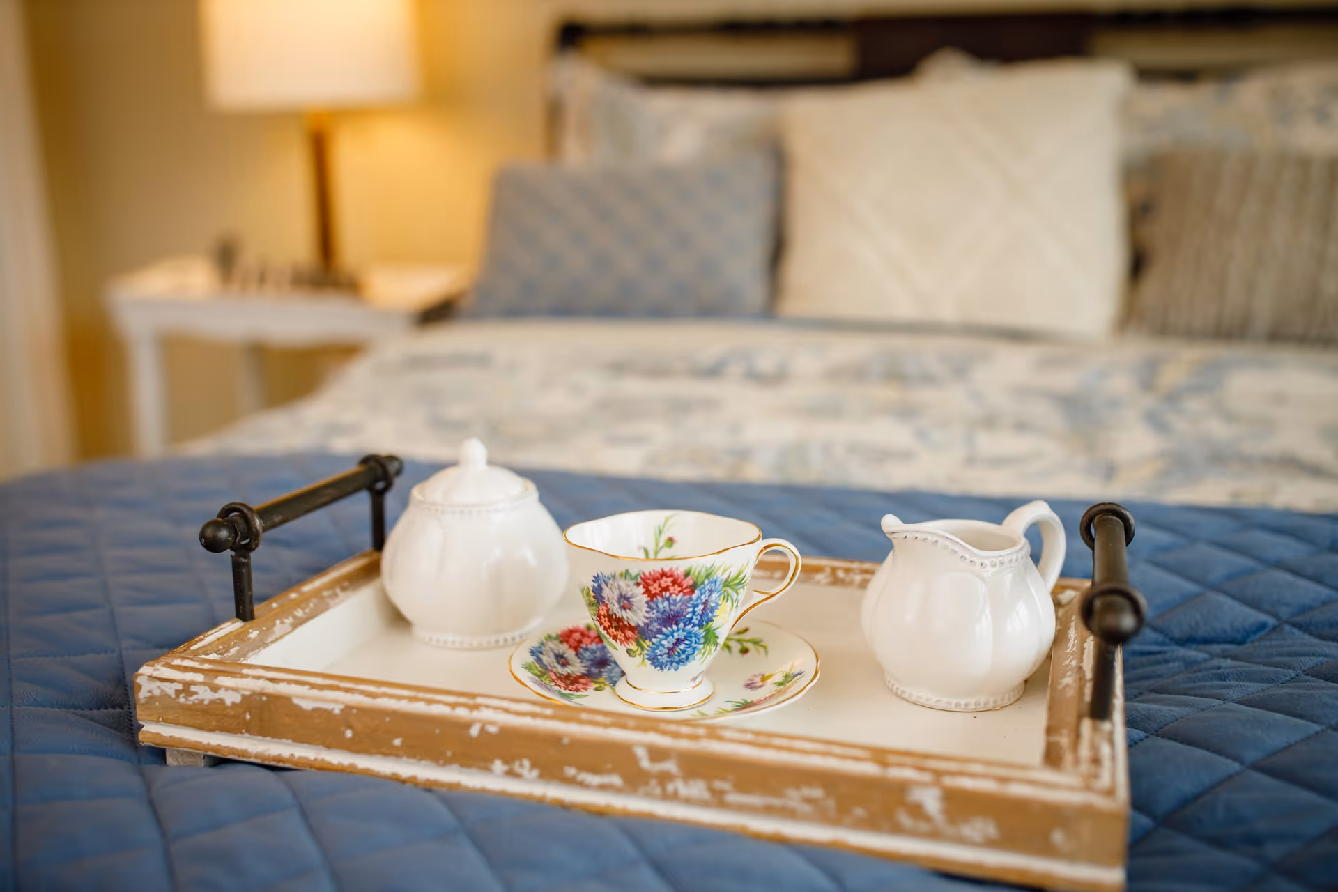 A wooden tray with black handles holding a floral teacup and saucer, a white sugar bowl with lid, and a white creamer pitcher, placed on a blue quilted bedspread with pillows and a lamp in the blurred background.