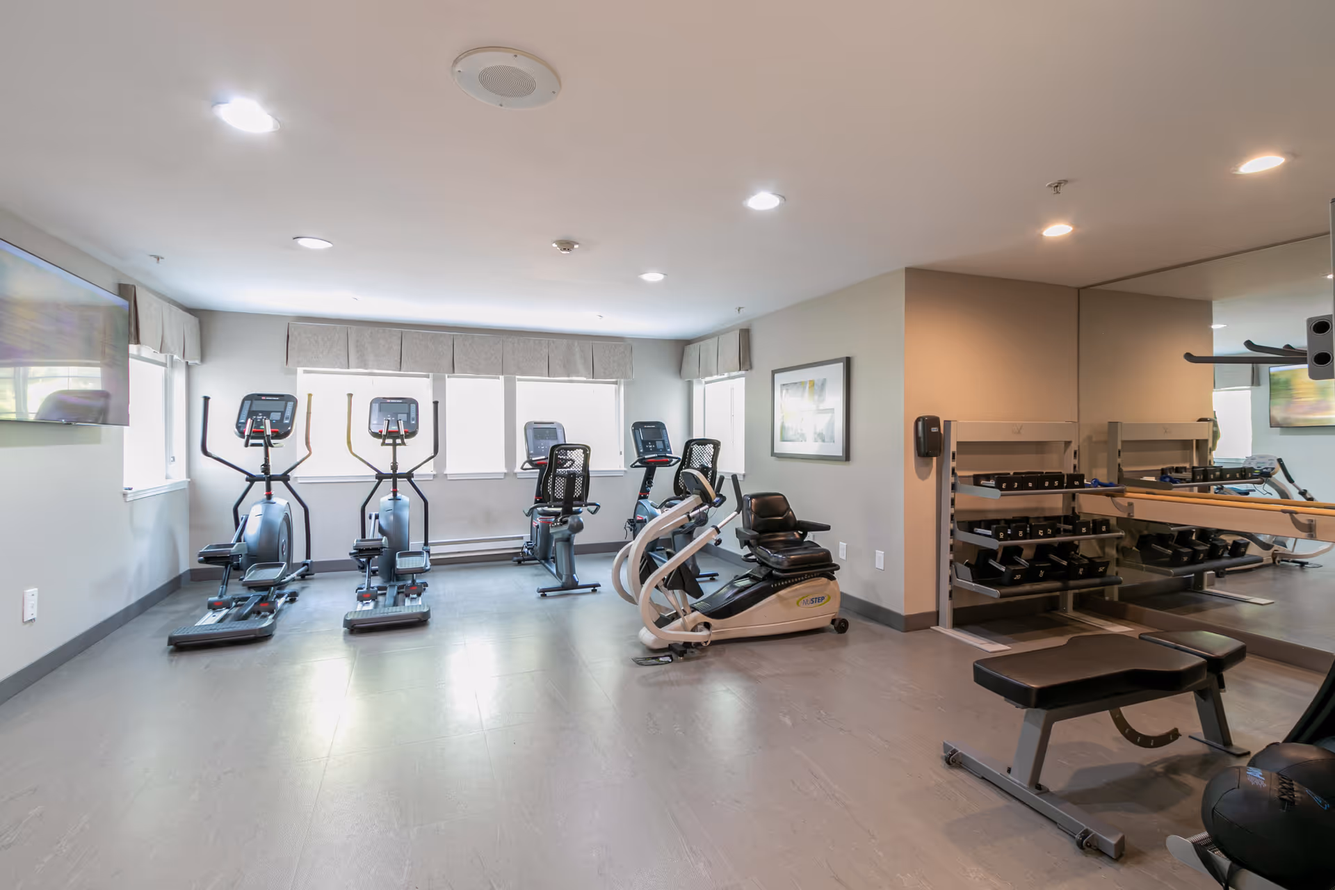A small fitness room with exercise equipment including stationary bikes, elliptical machines, a weight bench, and a rack of dumbbells. The room has large windows with valances, light gray walls, a wall-mounted TV, and a large mirror on one side.