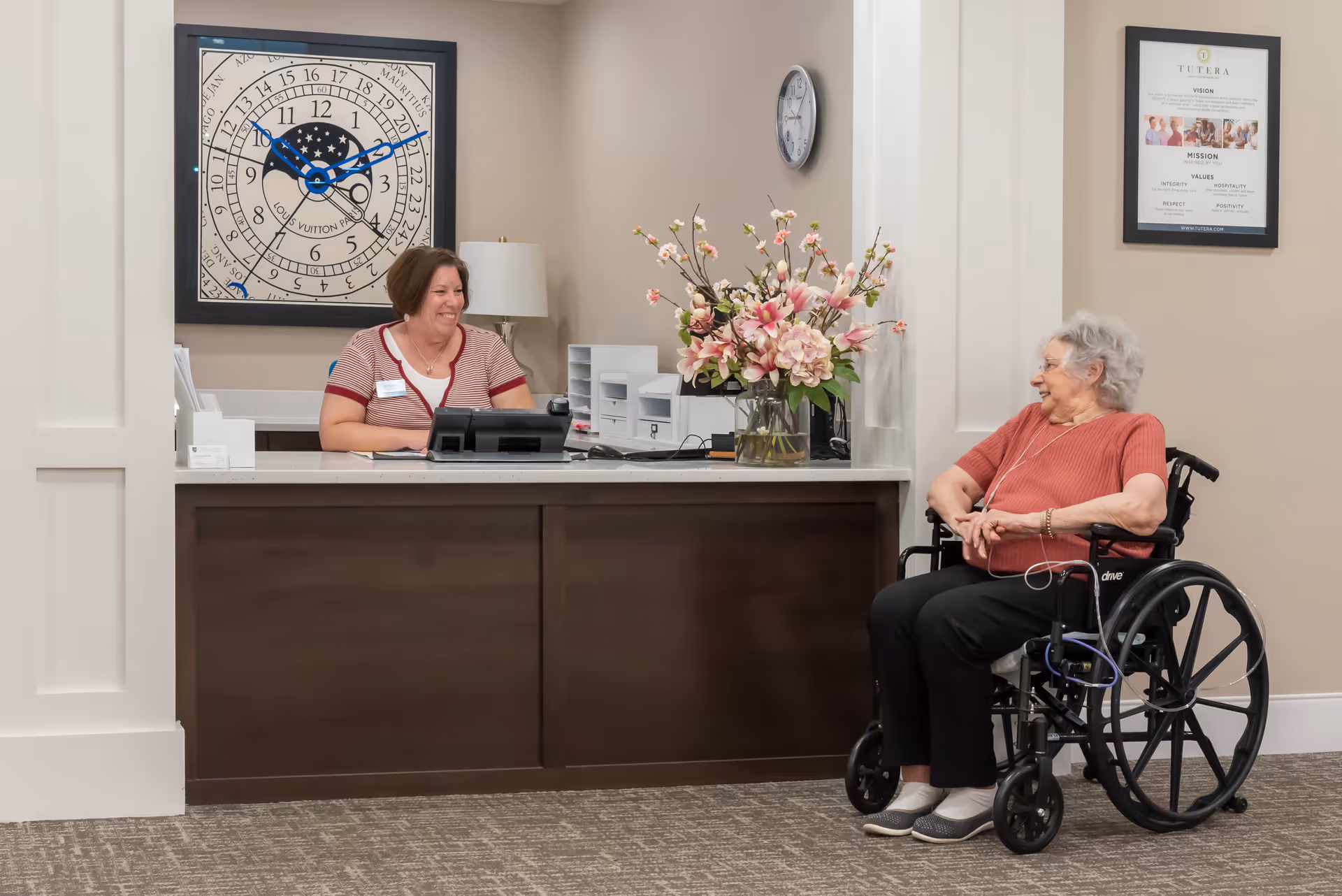 A smiling receptionist sitting behind a front desk talking to an elderly woman in a wheelchair. The desk has a large vase with pink flowers, a phone, and office supplies. Behind the receptionist is a large decorative clock on the wall, and a framed mission statement is visible on the wall to the right.