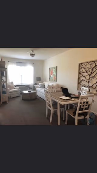 A cozy living room area with a white sofa, a round wooden coffee table, a white armchair near a window with sheer curtains, and a dining table with four white chairs. The room has neutral-colored walls with two pieces of wall art, one depicting flowers and the other a decorative tree design.